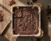 Overhead view of a wide rectangular image showing a square pan of baked vegan brownies with a crackly top and sea salt, on a rustic wooden table with chocolate and cocoa powder
